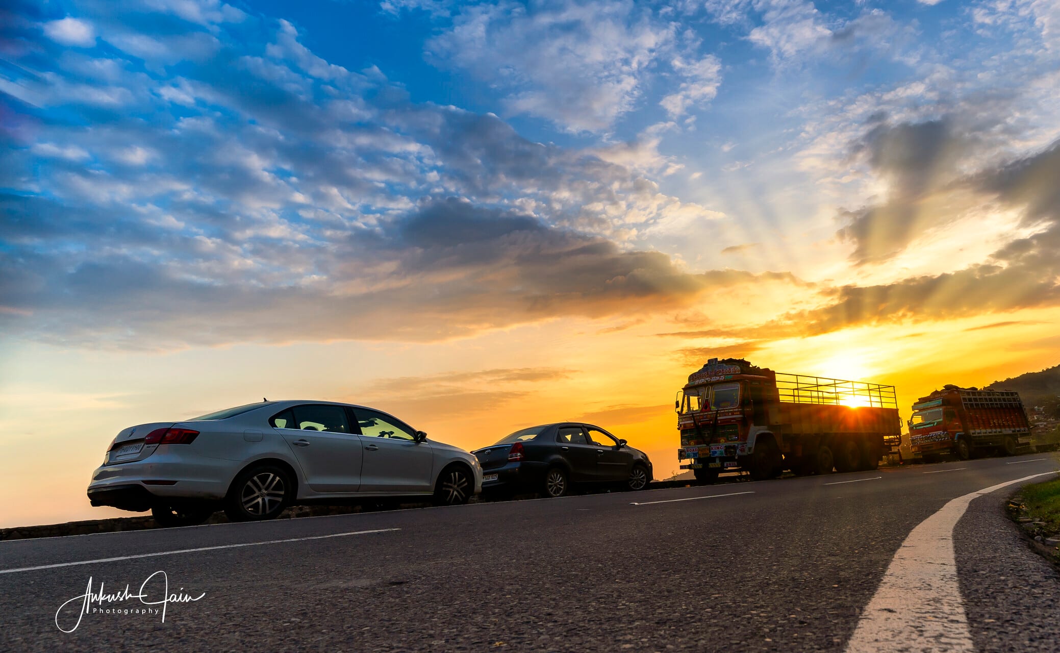 Cars parked at a hill-road overlook near Parwanoo, sun dropping behind a truck