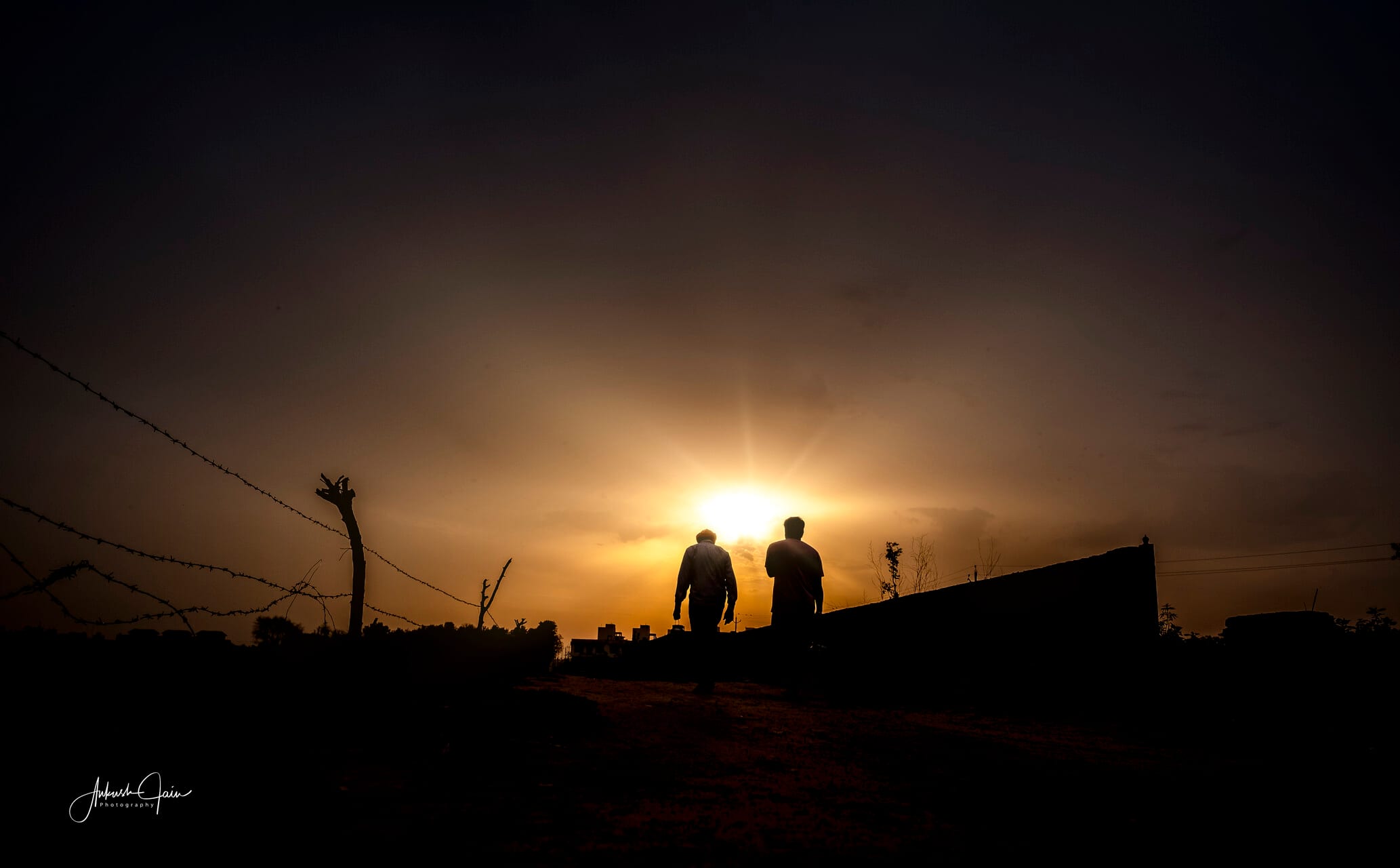 Two figures walking toward the rising sun past a barbed-wire fence, village Barewal