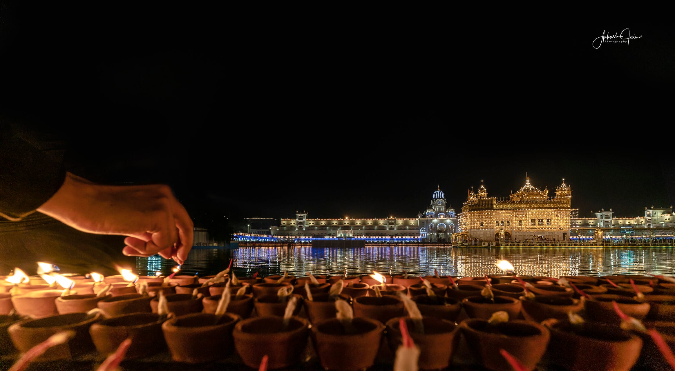 A hand lighting rows of clay diyas beside the lit temple on Gurpurab night