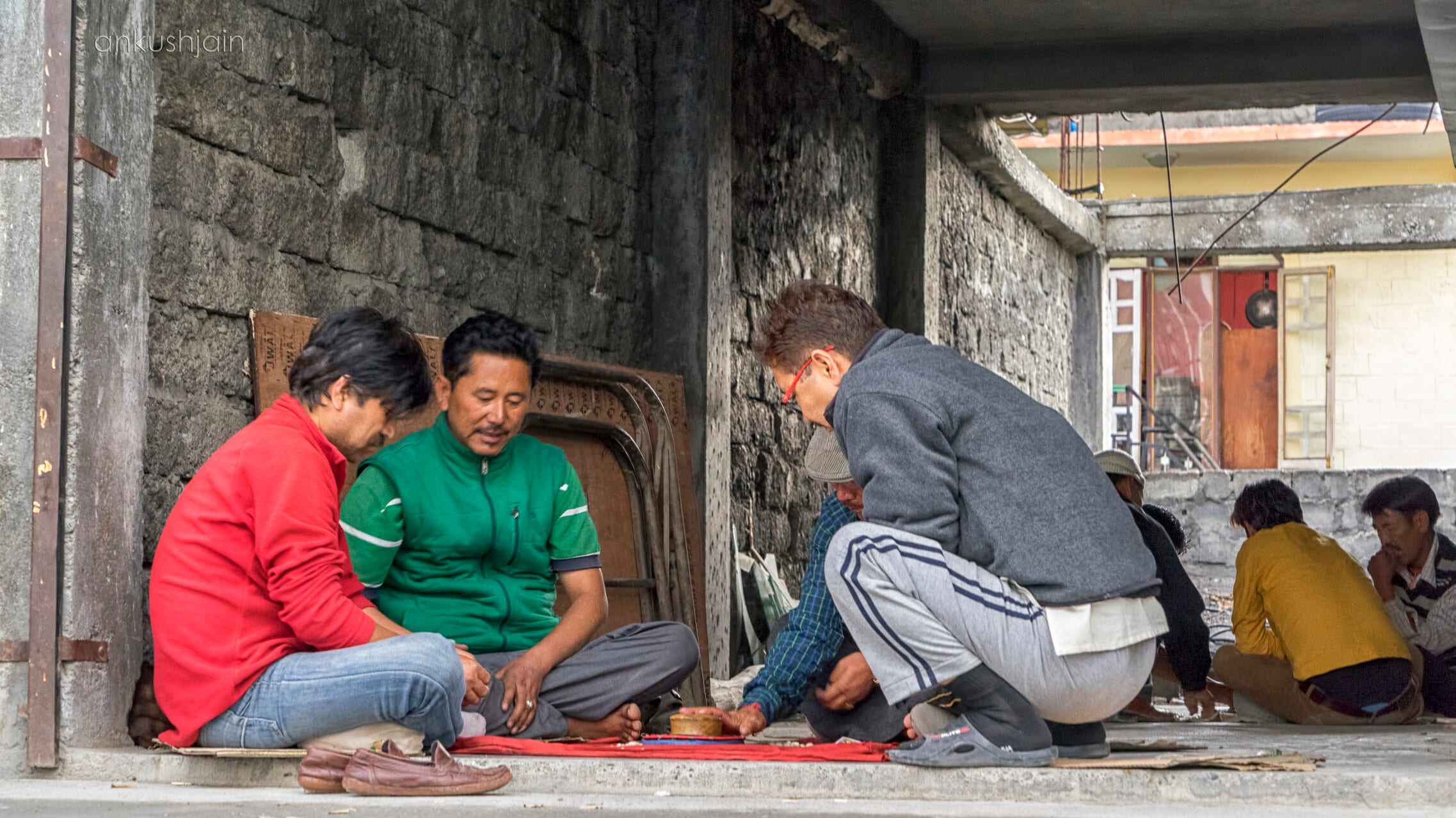 Three men hunched over a board game on a stone verandah in Spiti, onlookers resting beside them