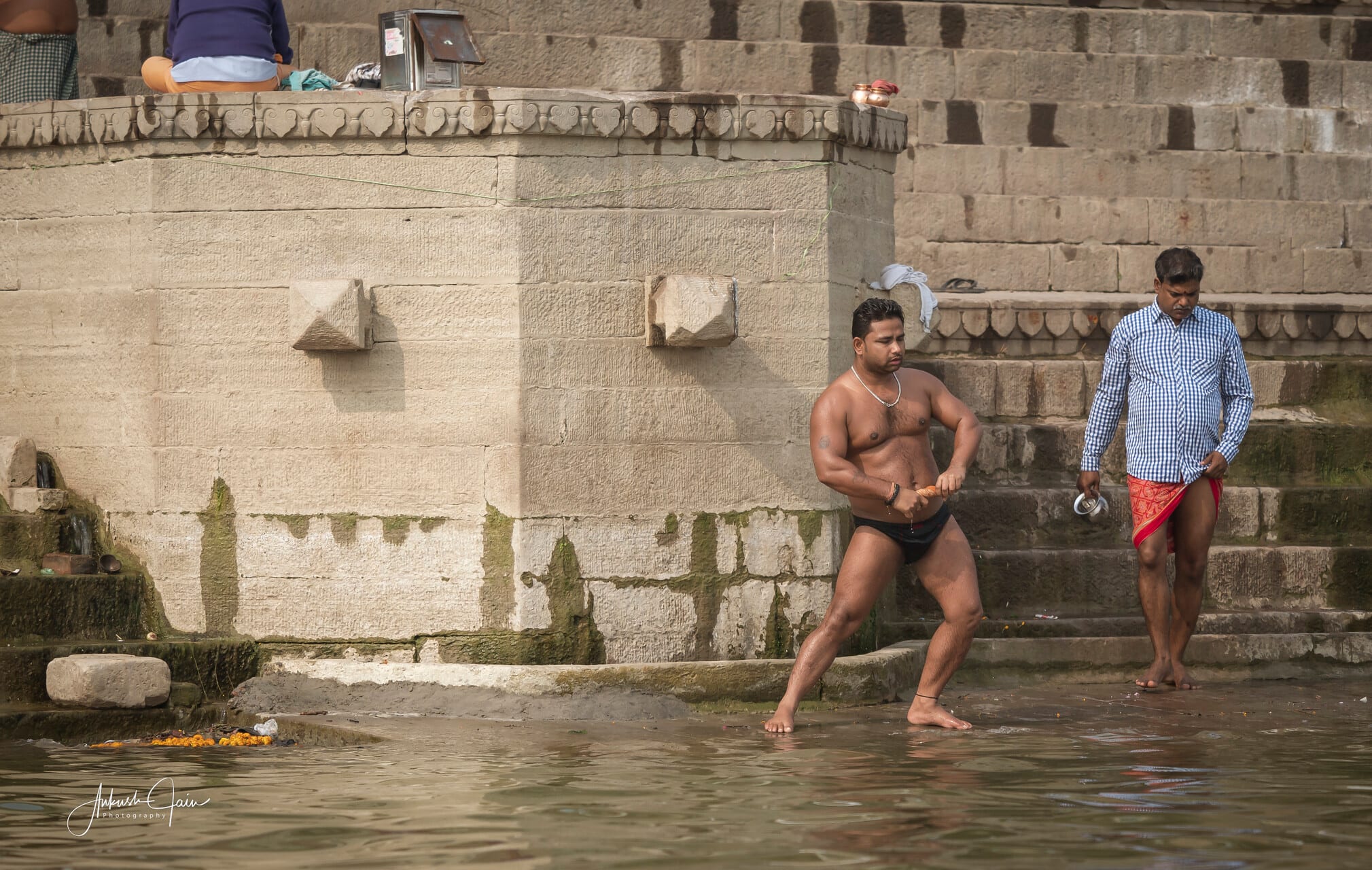 Bodybuilder flexing at a stone ghat in Varanasi, marigold offerings floating on the water