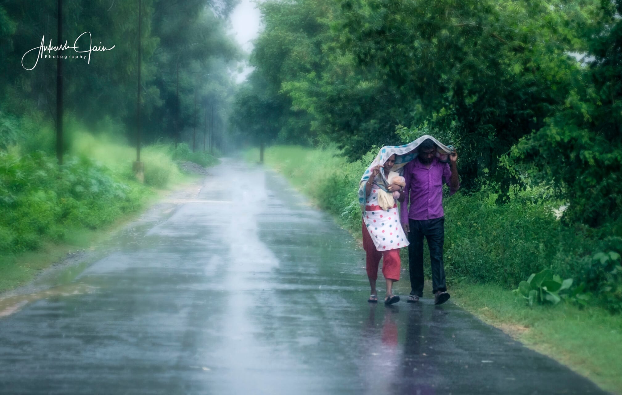 Couple sheltering under a cloth on rainy Sua Road near Barewal, woman carrying a baby