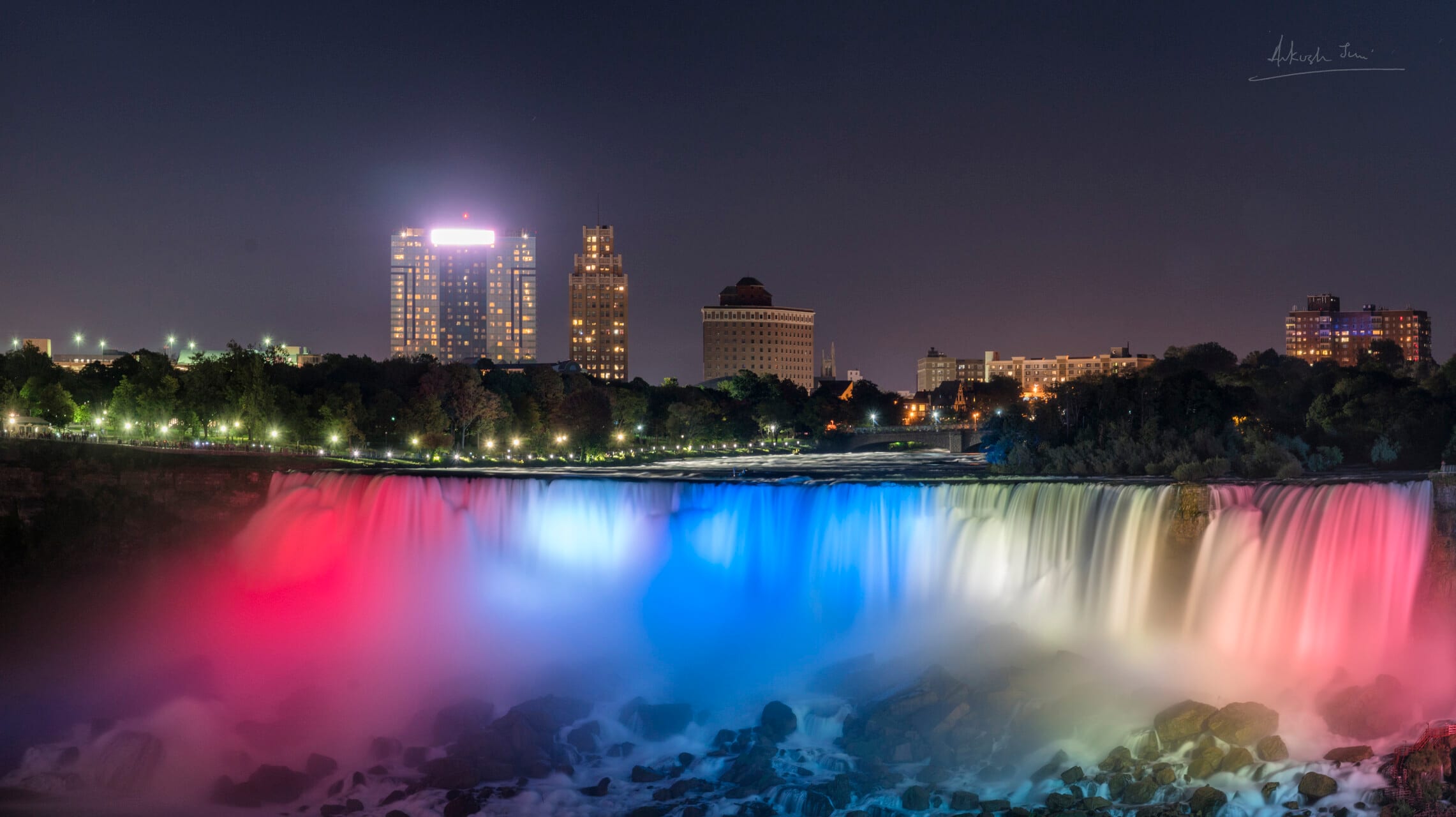 Niagara Falls lit red, white and blue at night, the city skyline glowing behind