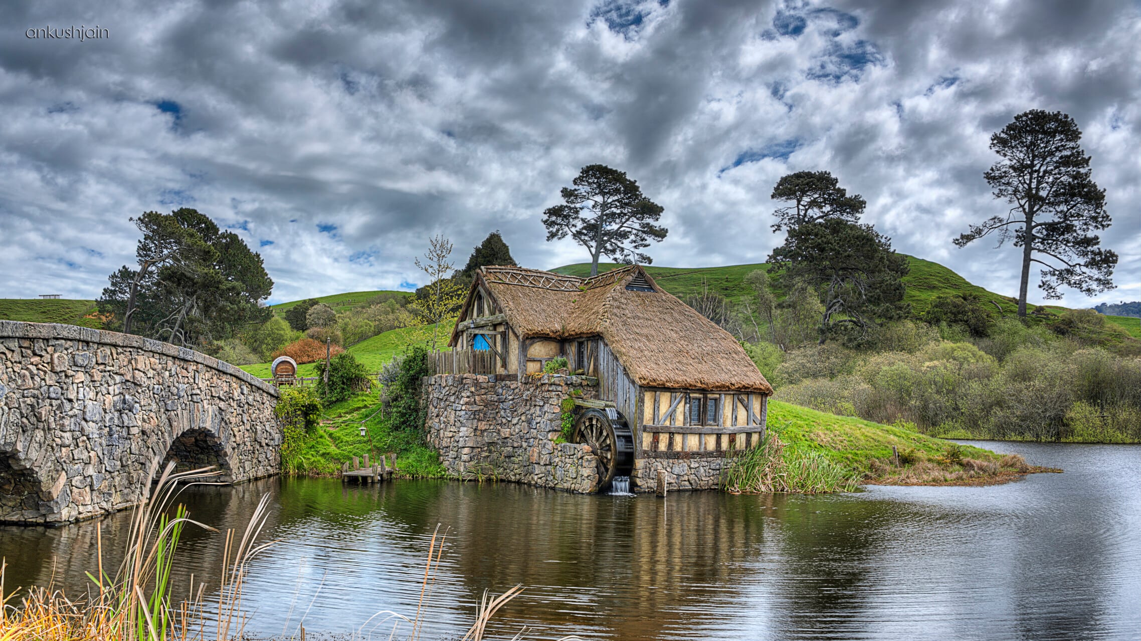 The Mill cottage and stone bridge at Hobbiton, thatched roof and waterwheel mirrored in the pond