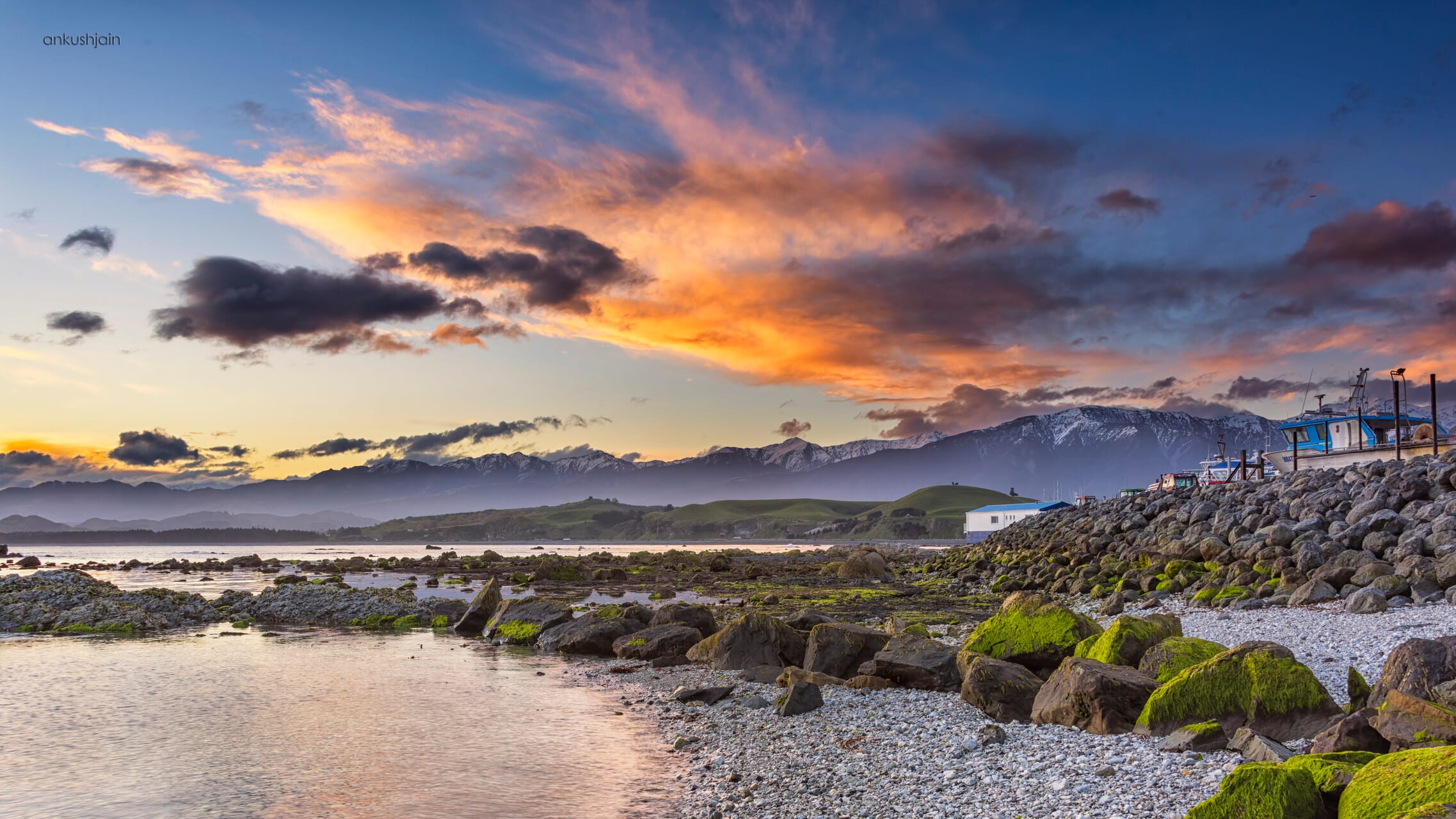 Sunset over the Kaikoura coast, mossy boulders in the foreground and snow peaks beyond