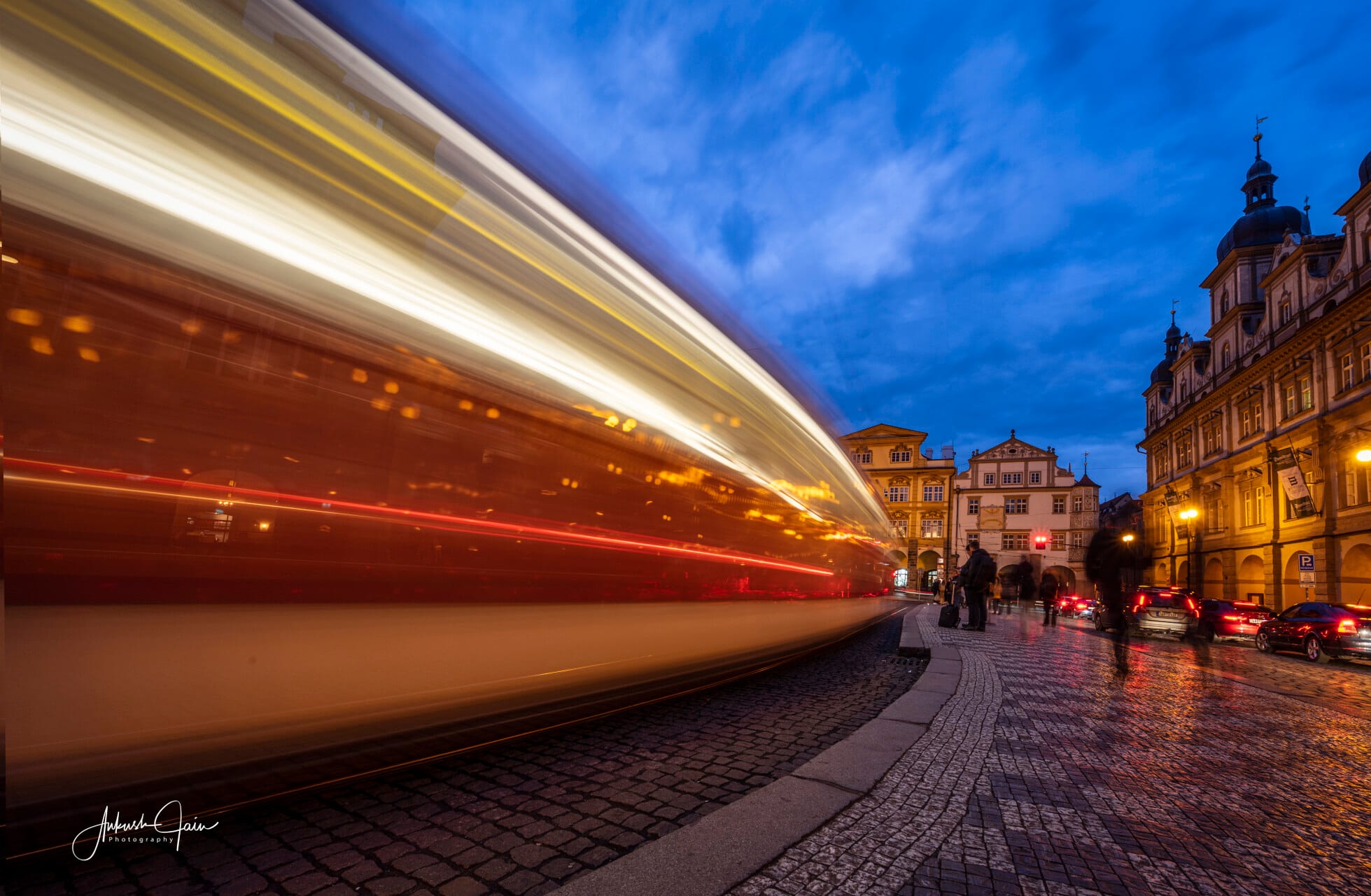 Tram light-trail rushing through Malá Strana at blue hour, baroque facades lining the cobblestone street
