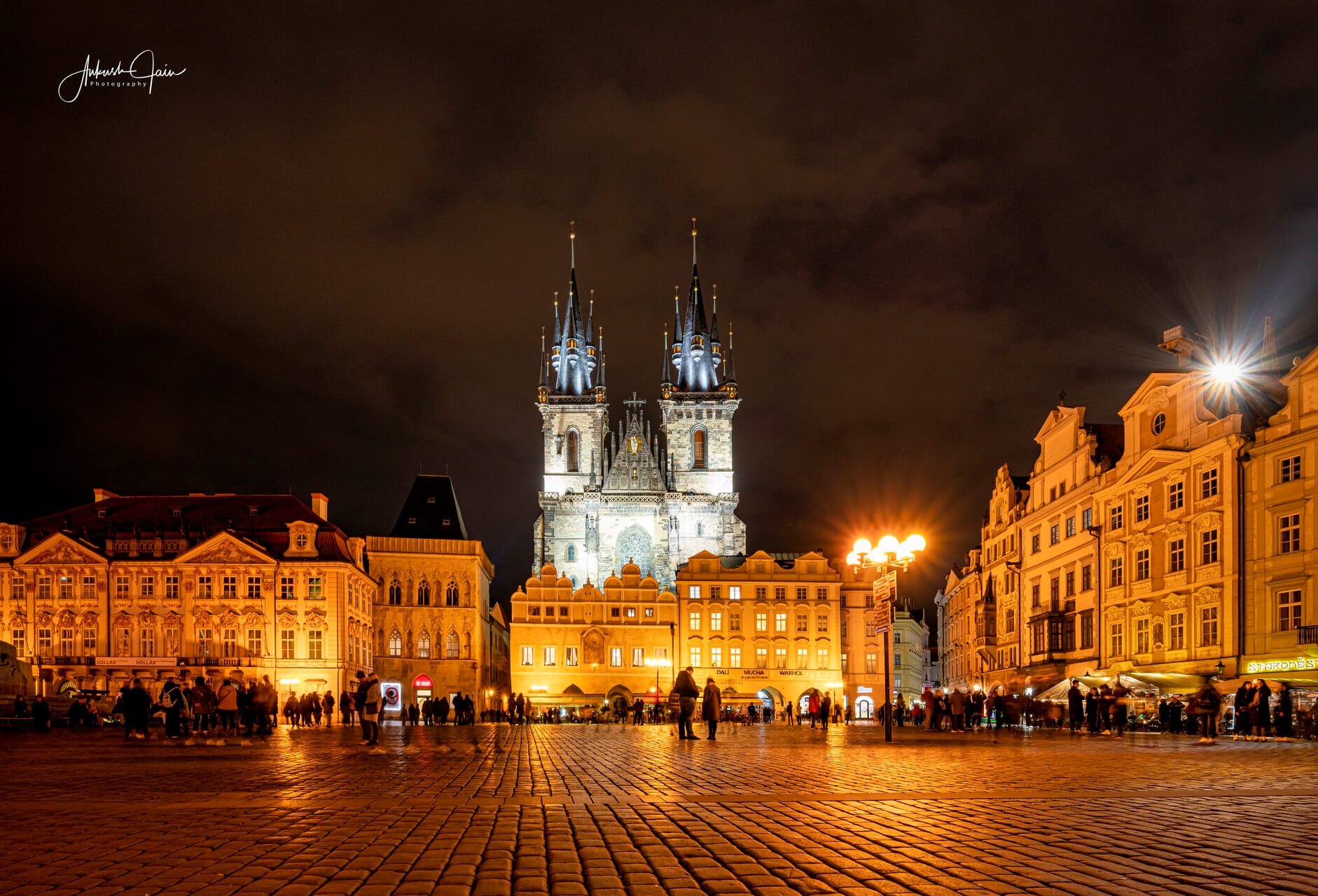 Prague’s Old Town Square at night — the twin Gothic spires of the Týn Church rising behind the warmly lit baroque facades, cobblestones glowing under the lamps