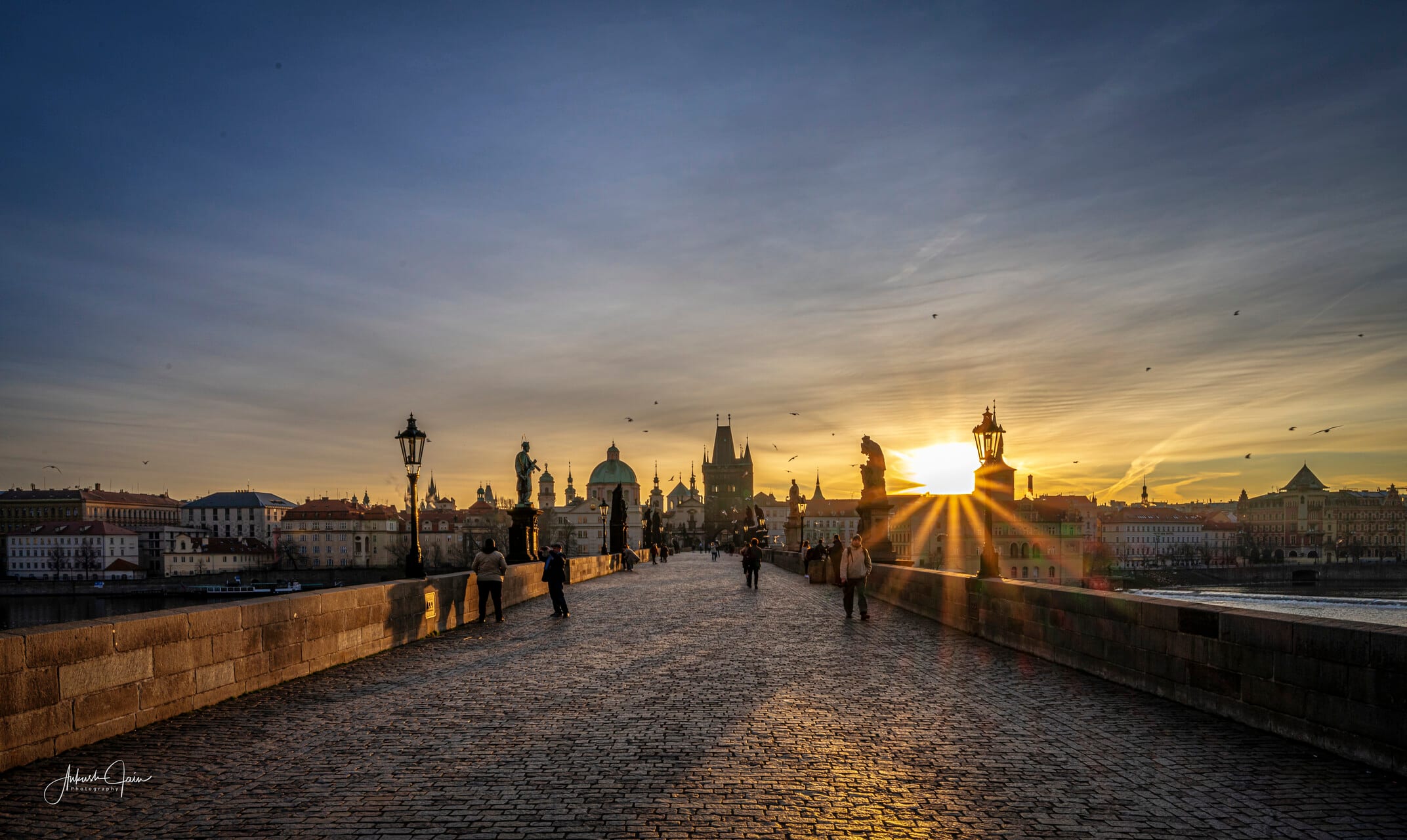 Sunrise from Charles Bridge, lampposts and the Old Town skyline silhouetted against the rising sun