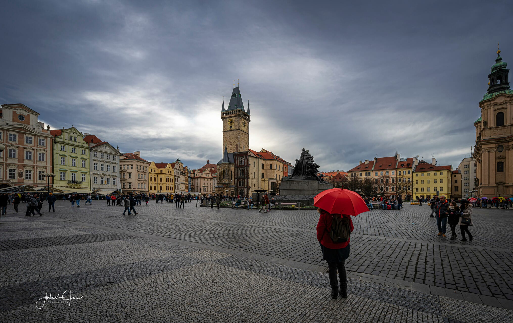 Lone figure with a red umbrella in Old Town Square, the Old Town Hall tower rising under a grey sky