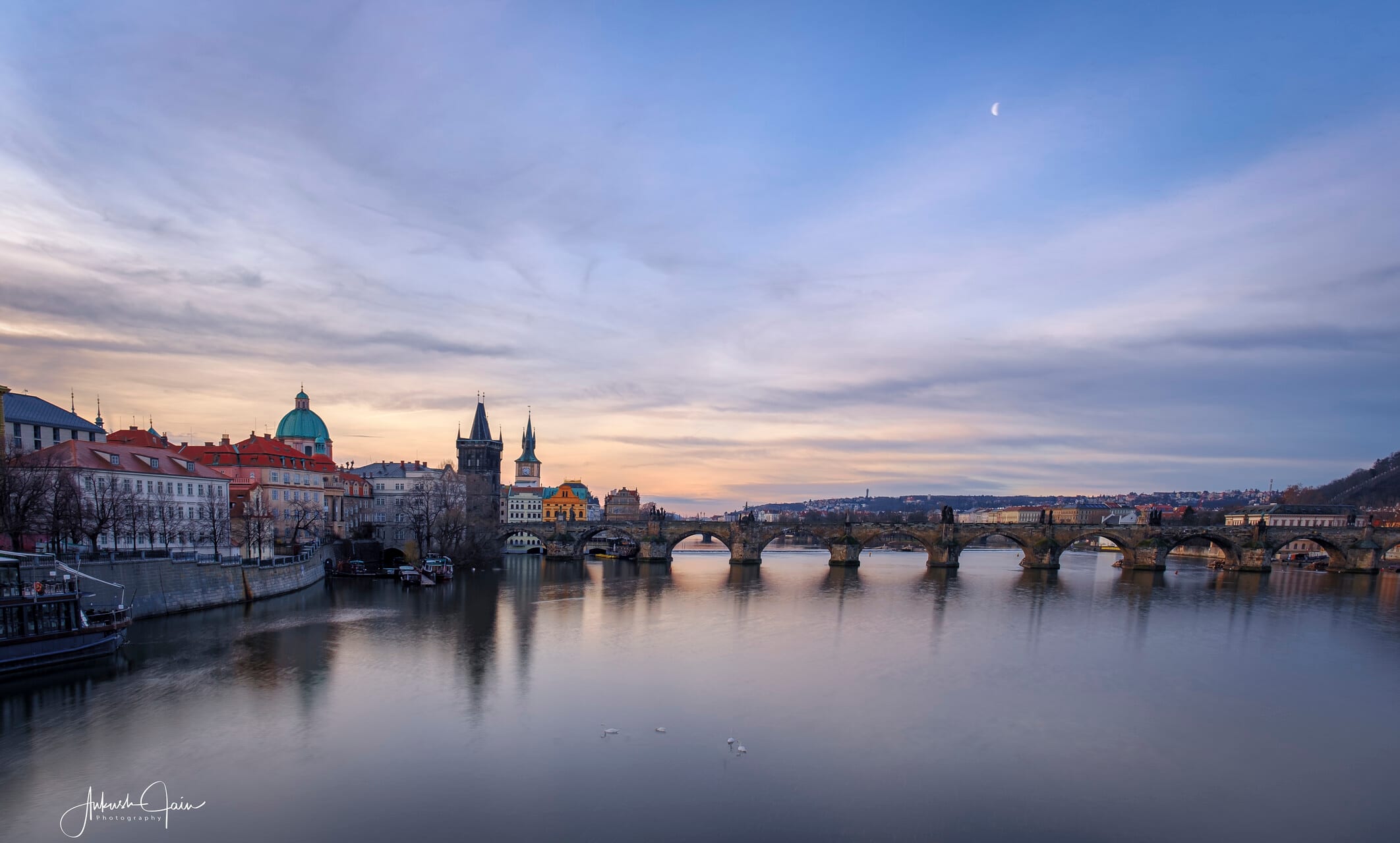 Charles Bridge and the Old Town spires from across the Vltava at dusk, crescent moon rising overhead