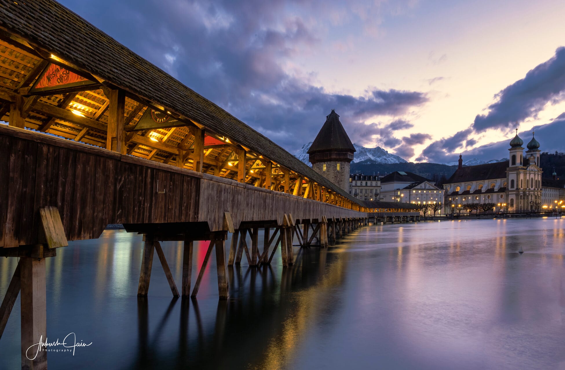 Lucerne’s Chapel Bridge at blue hour — the covered wooden walkway and octagonal water tower lit against an overcast sky, the Jesuit Church glowing across the Reuss