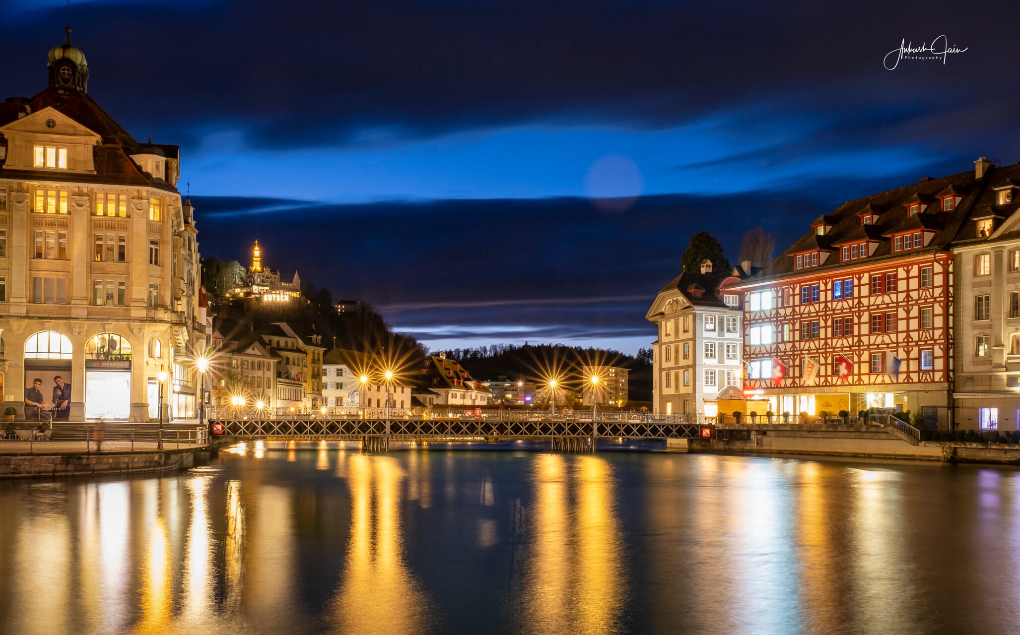 Lucerne at night — half-timbered facades on one bank, Château Gütsch lit on the hill across the river, street lamps starring on the still water