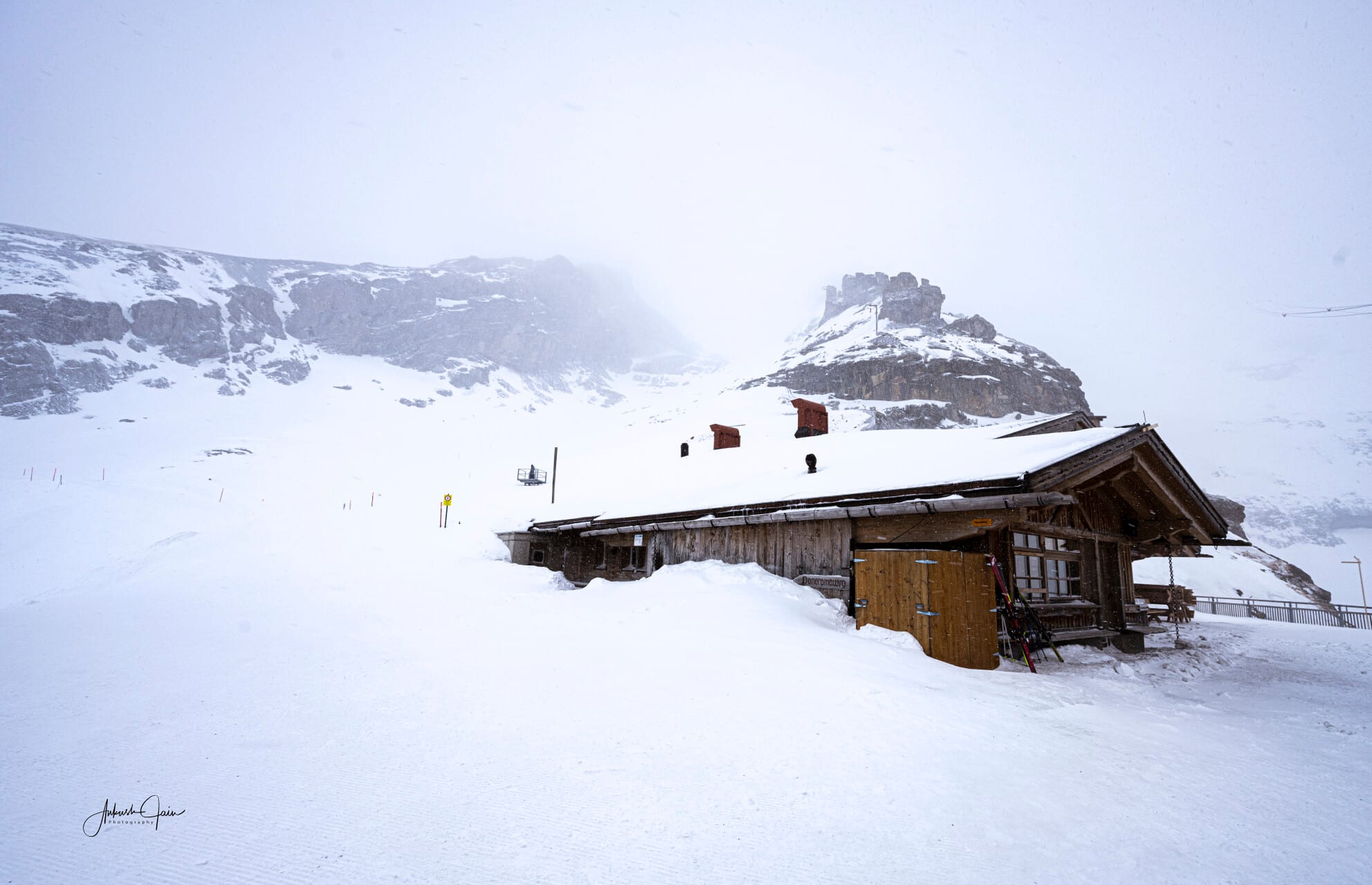A weathered wooden ski hut half-buried in fresh snow on a Jungfrau slope, rock and cloud fading into white behind it
