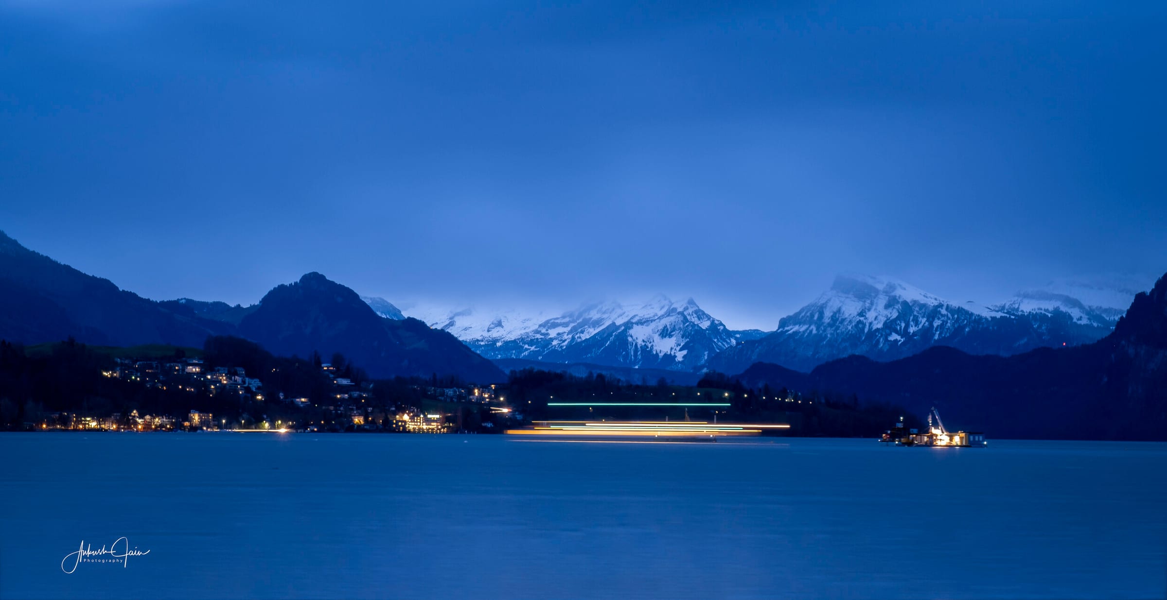 Long exposure of a boat crossing Lake Lucerne at blue hour, its light trail stretched across the water with the snow-dusted Alps behind