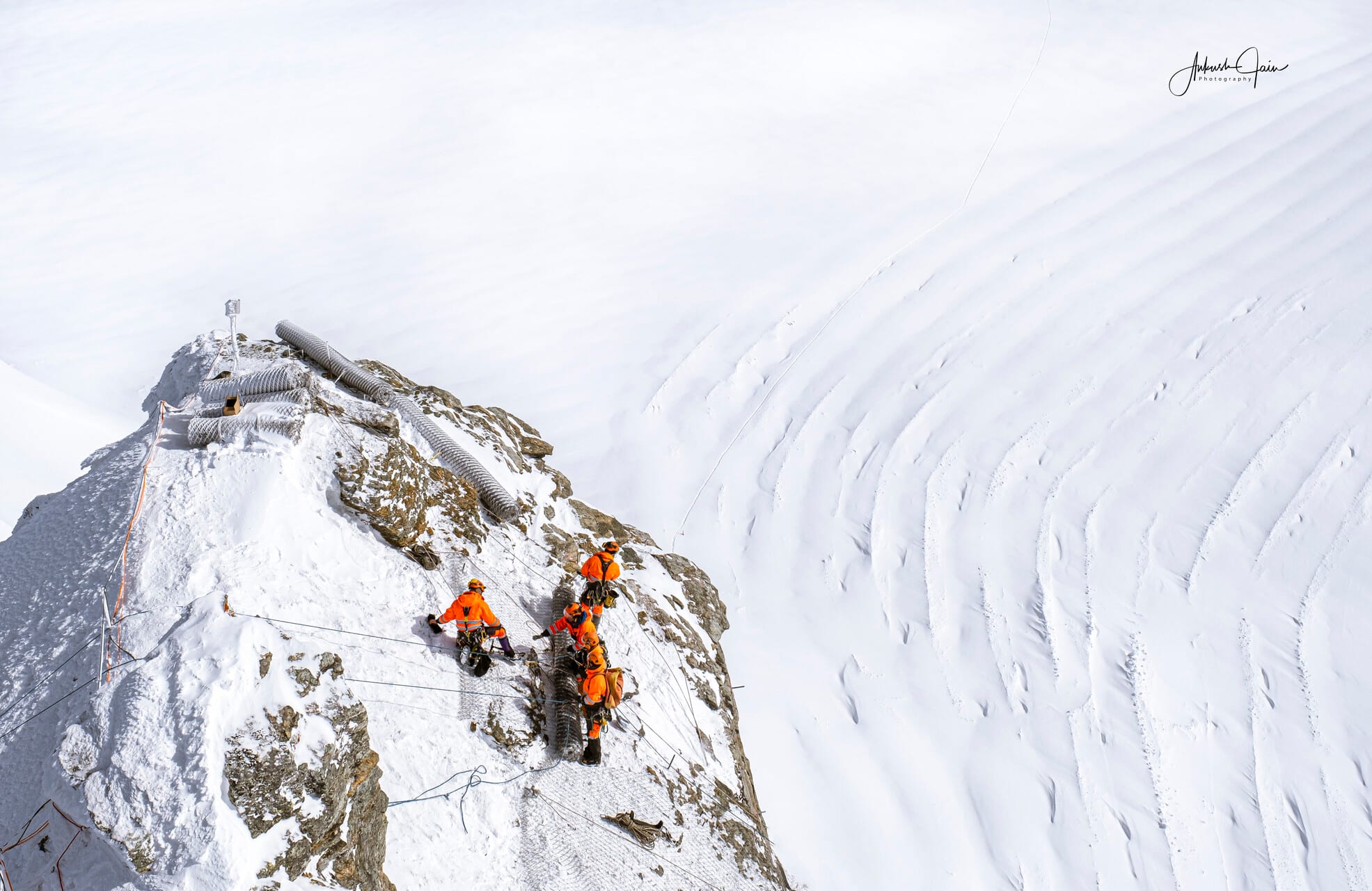 Workers in orange suits roped onto a rocky outcrop near the Jungfraujoch summit, the wind-sculpted snow slope falling away to the right