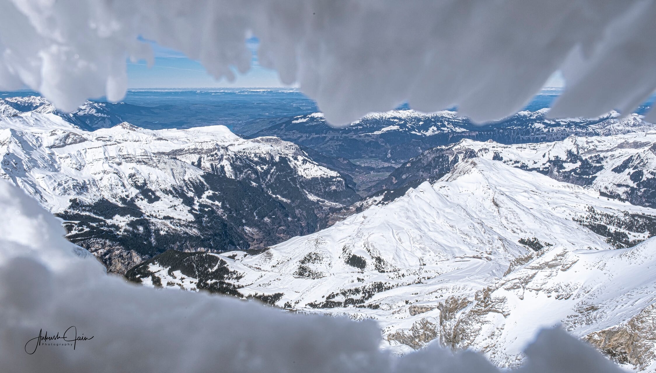 Panorama of snowy peaks and a valley far below, framed by icicles hanging from a high-altitude vantage