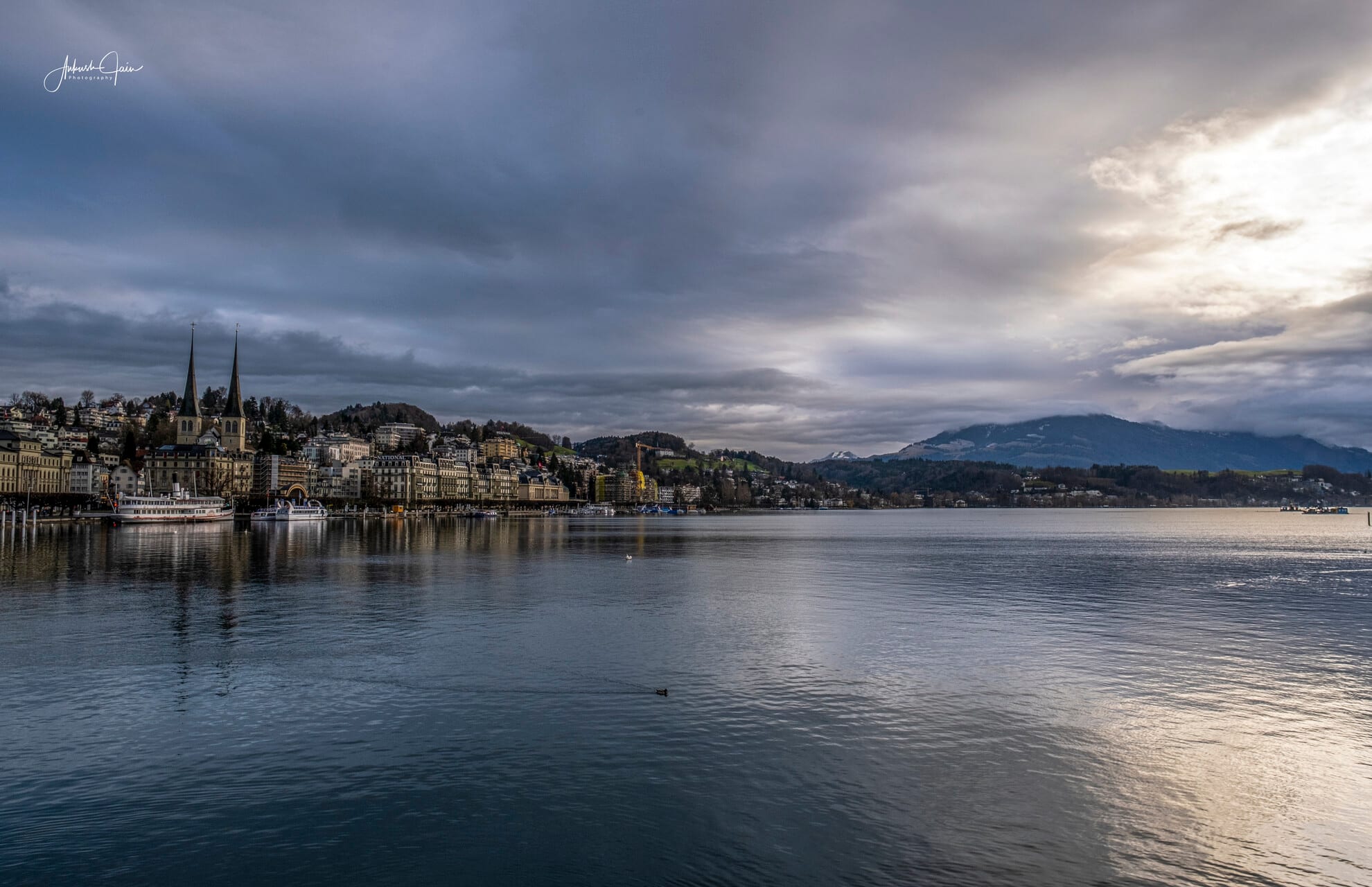 Lake Lucerne under a moody late-afternoon sky — the twin spires of the Hofkirche and a paddle steamer at the dock on the far bank
