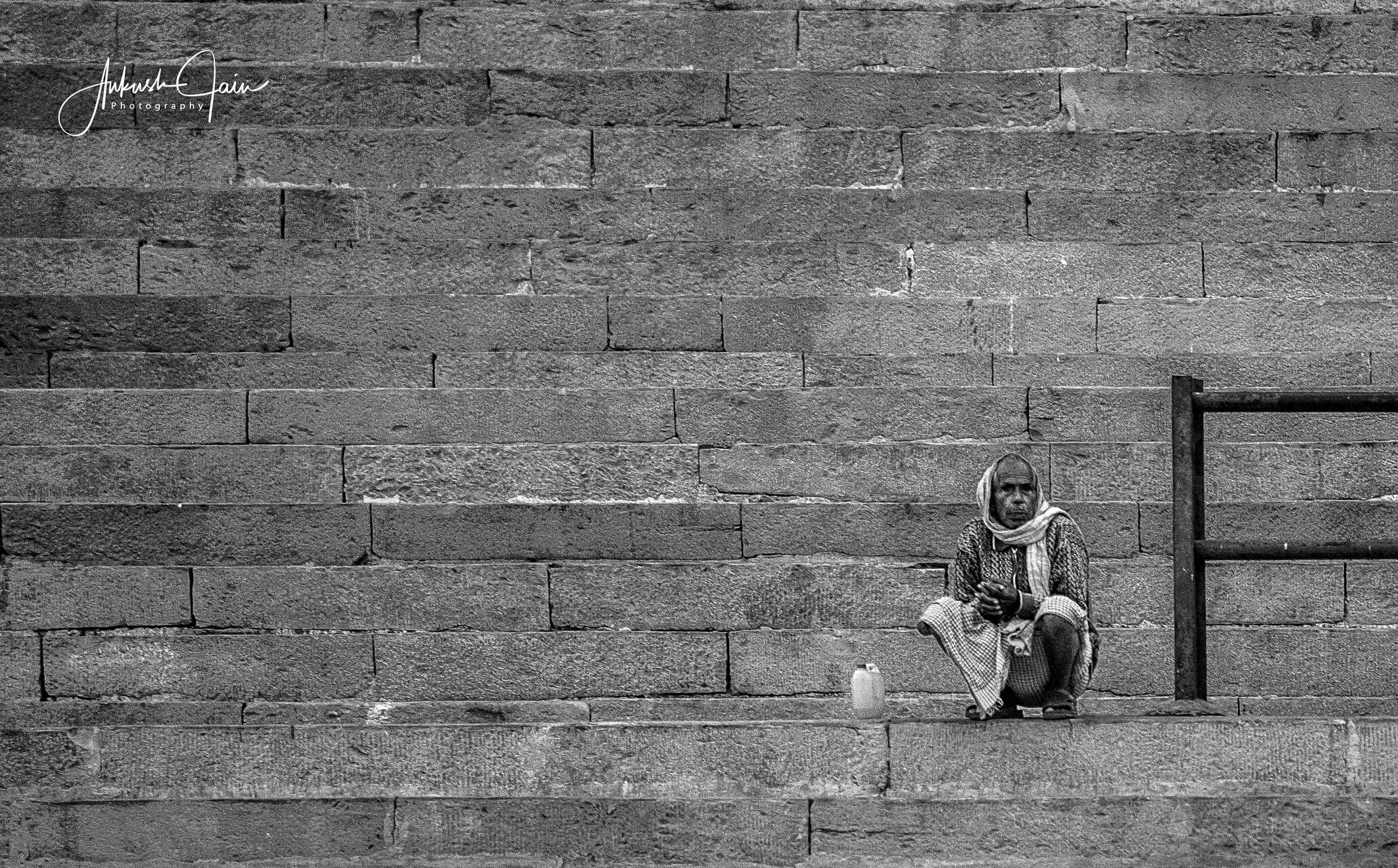 Black-and-white of an elderly man sitting small against the layered stone wall of a Varanasi ghat, a tin cup beside him