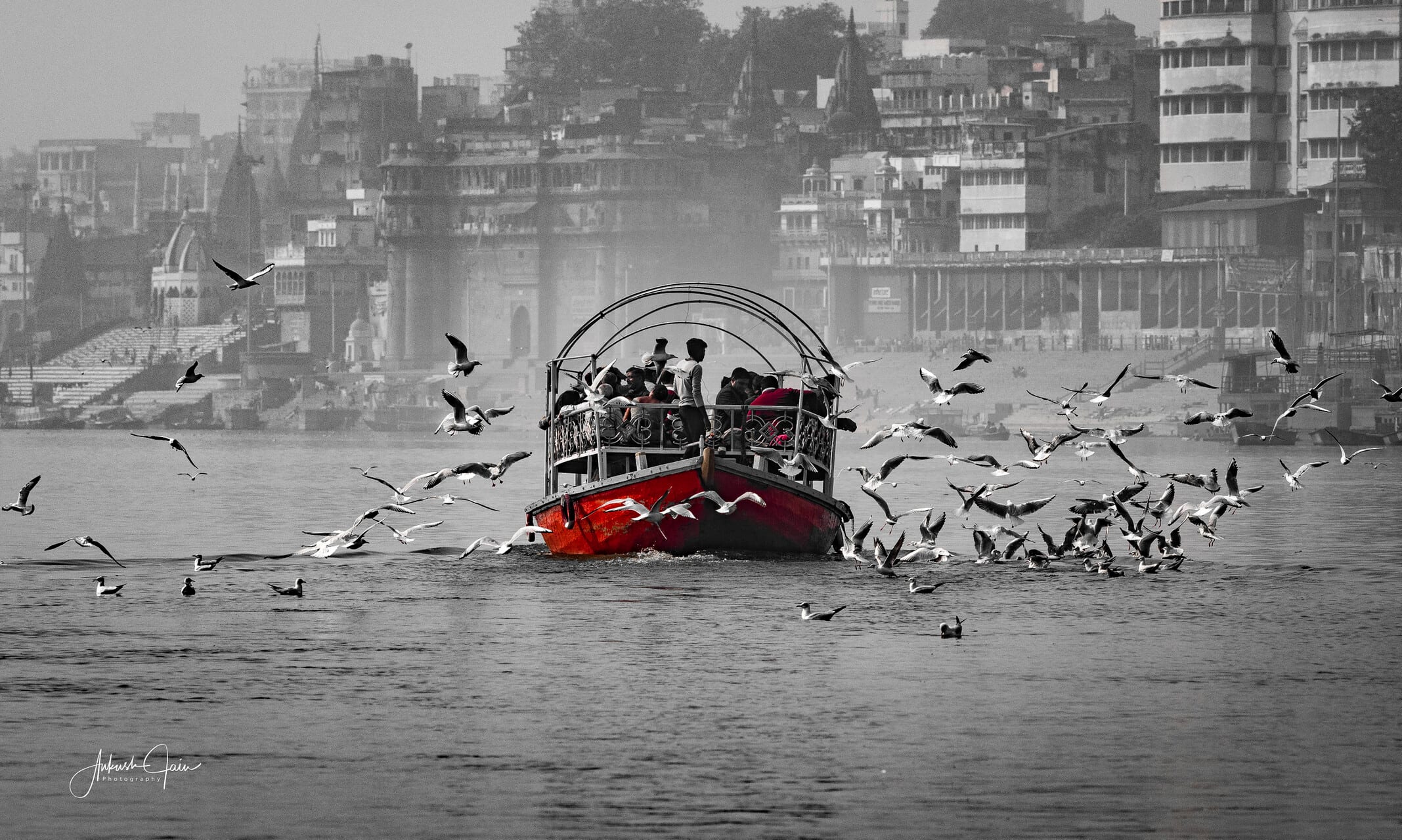 Selective colour: a red boat on the Ganga as seagulls swirl around it, ghats receding in monochrome behind