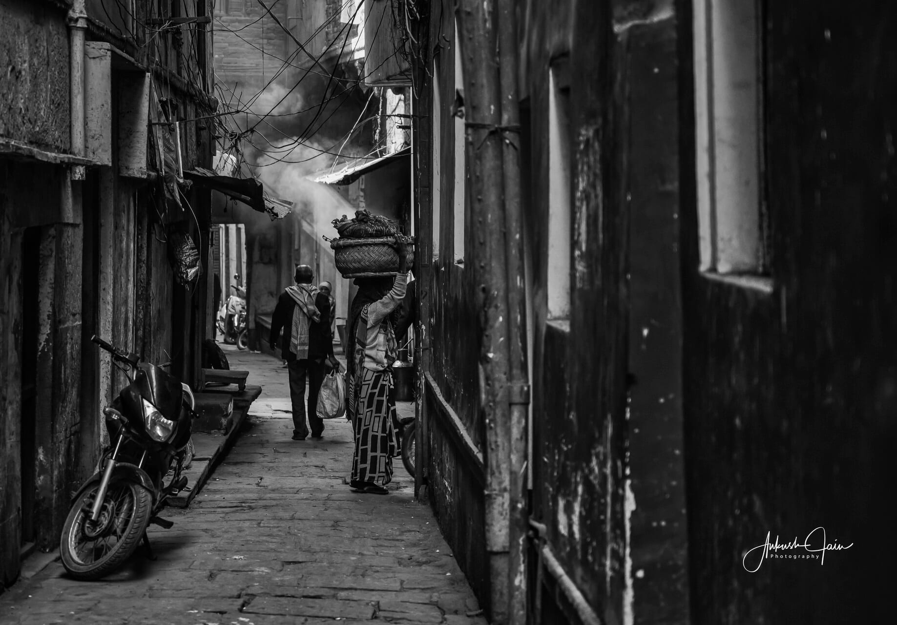 Black-and-white of a woman carrying a basket on her head through a narrow Varanasi alley, light shaft cutting the smoke