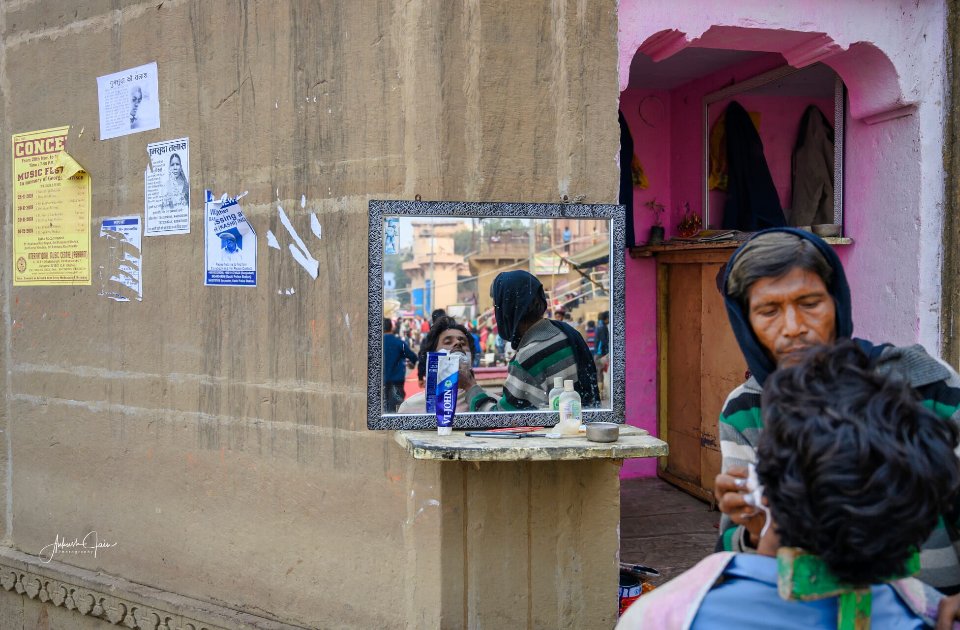 A wall-mounted mirror at an open-air barber stall in Varanasi reflects a customer mid-shave with the ghat crowds behind, while missing-person flyers and a Music Festival poster paper the wall beside it