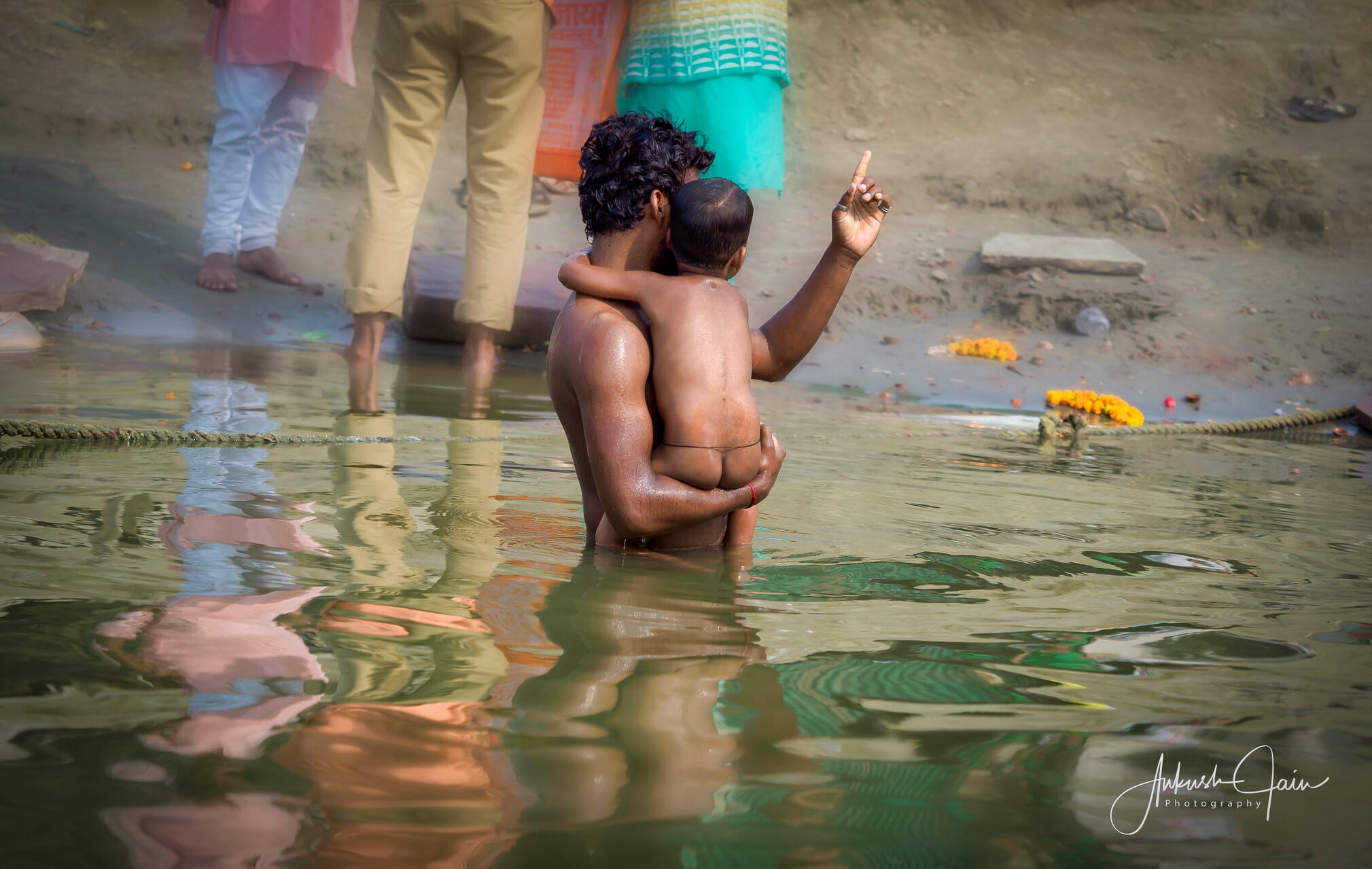 A father holding his small son for a holy dip in the Ganga, marigold offerings floating beside them