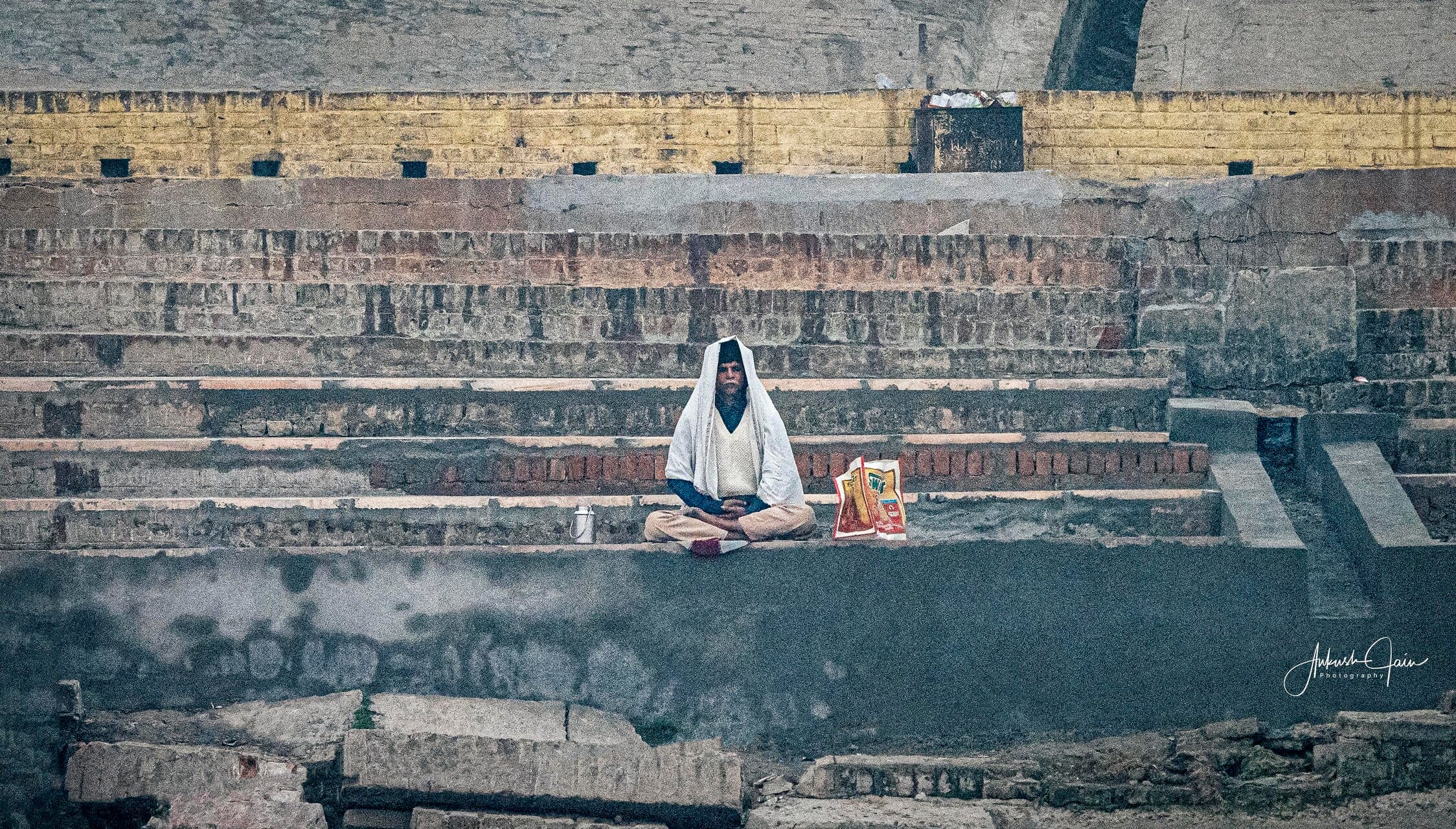 A sadhu in a white shawl meditating on a stone step at the ghat, offerings beside him