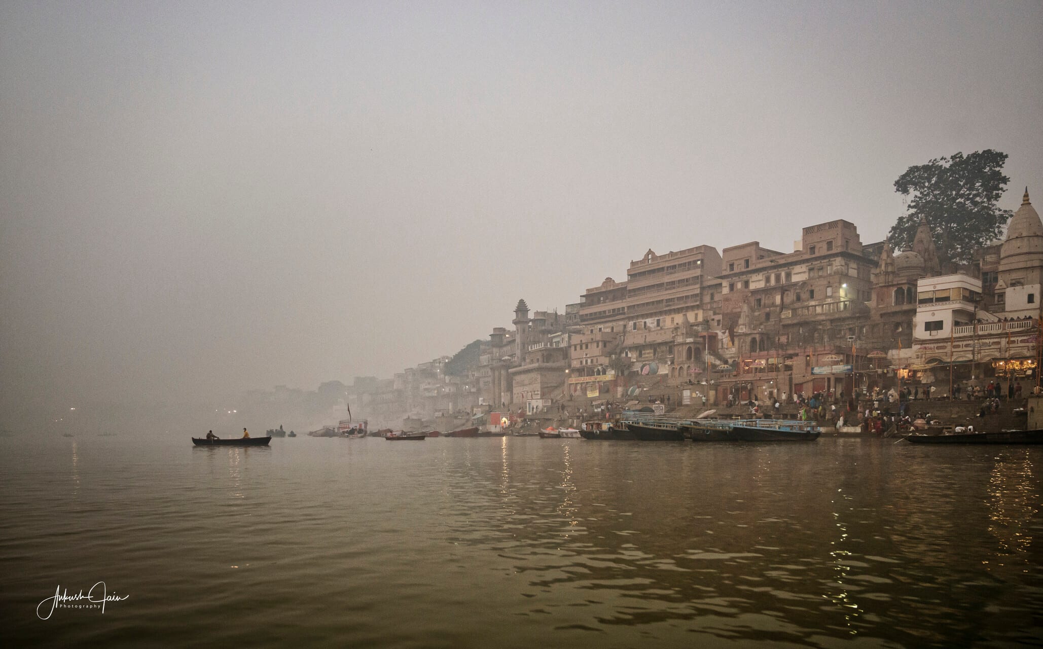 Wide misty morning view of the Varanasi ghats, a lone rowboat drifting in the foreground